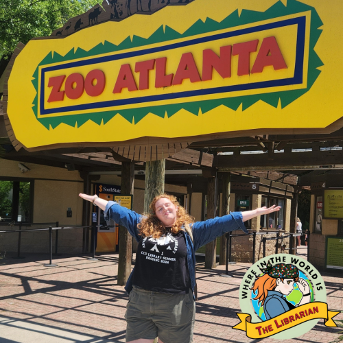 A red-haired woman in a black t-shirt, denim overshirt, and green shorts standing with her arms outstretched in front of the Zoo Atlanta sign.