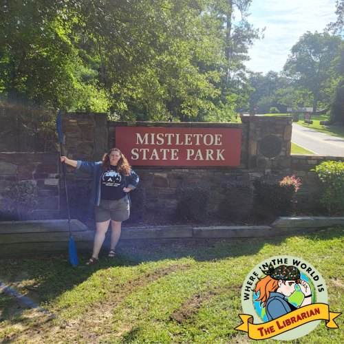 A red-haired woman in a black t-shirt and green shorts in front of the Mstletoe State Park sign