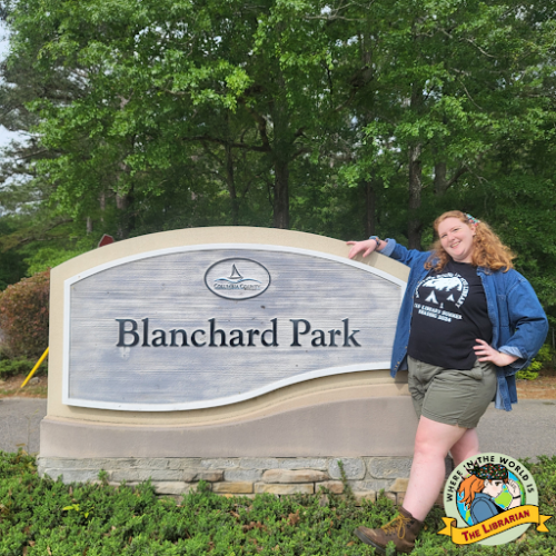 A red-haired woman in a black t-shirt, green shorts, brown hiking boots, and a kerchief standing next to the entrance sign for Blanchard Park